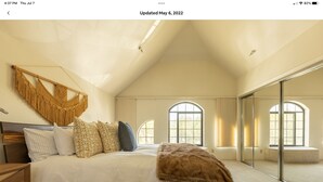 Un espacioso dormitorio con una cama grande, un cabecero de madera, una decoración de pared colgante de macramé y ventanas amplias.