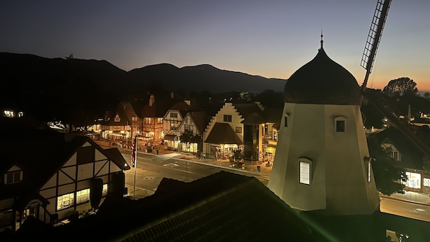 Un molino de viento tradicional y una hilera de casas con entramado de madera y ventanas iluminadas, todo sobre un fondo de colinas.