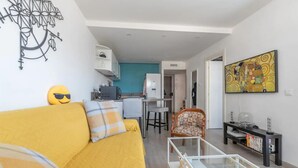 Ein Wohnzimmer mit einem gelben Sofa, einem Glastisch und einer Küchenzeile, die einen weißen Kühlschrank und eine blaue Wand beinhaltet.