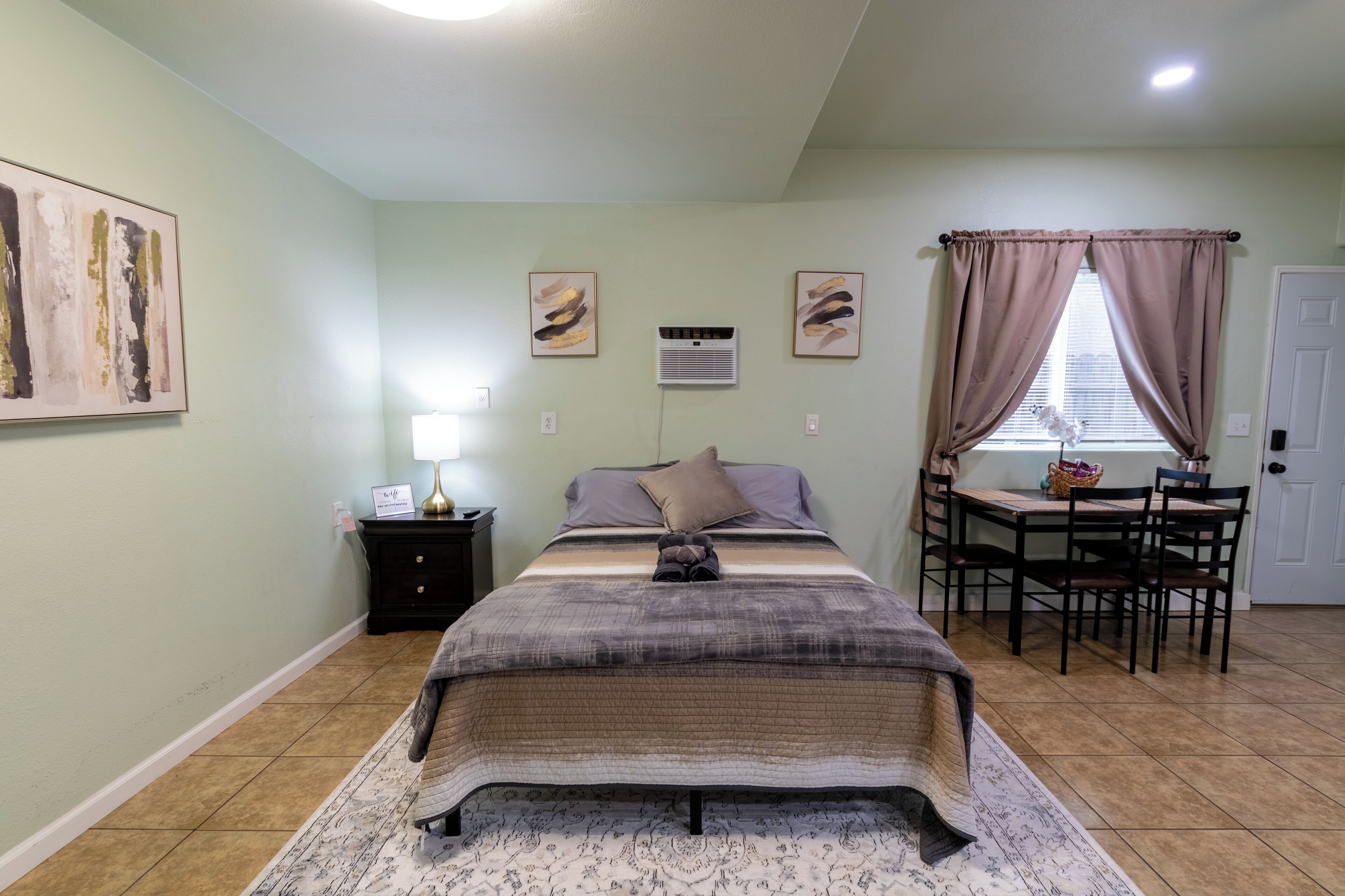 Un dormitorio con cama, mesita de noche, lámpara, equipo de aire acondicionado, ventana con cortinas y una mesa de comedor con sillas.