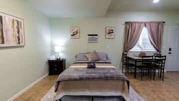 Un dormitorio con cama, mesita de noche, lámpara, equipo de aire acondicionado, ventana con cortinas y una mesa de comedor con sillas.