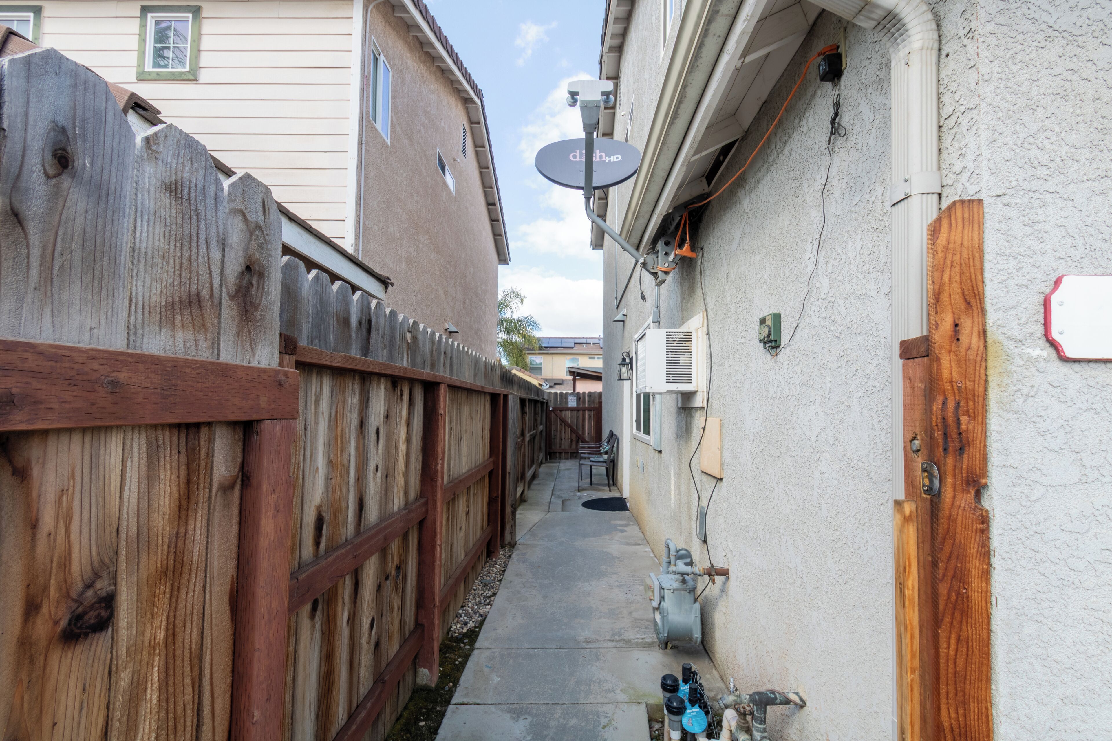 A narrow alleyway with a wooden fence, a concrete path, and a building with a satellite dish and utility boxes.