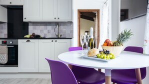 Una cucina moderna con mobili bianchi, un tavolo da pranzo viola e sedie. Sul tavolo c'è una ciotola di frutta e una bottiglia di vino.