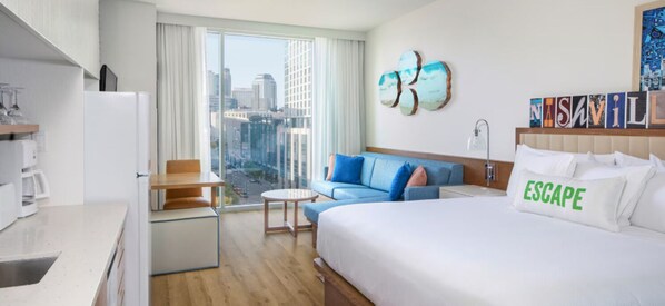 A hotel room with a bed, a sofa, a small table, a kitchen area, and a large window with a city view.