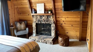 Una habitación acogedora con chimenea de piedra, un televisor montado en la pared, un sillón cómodo y un banco acolchado.