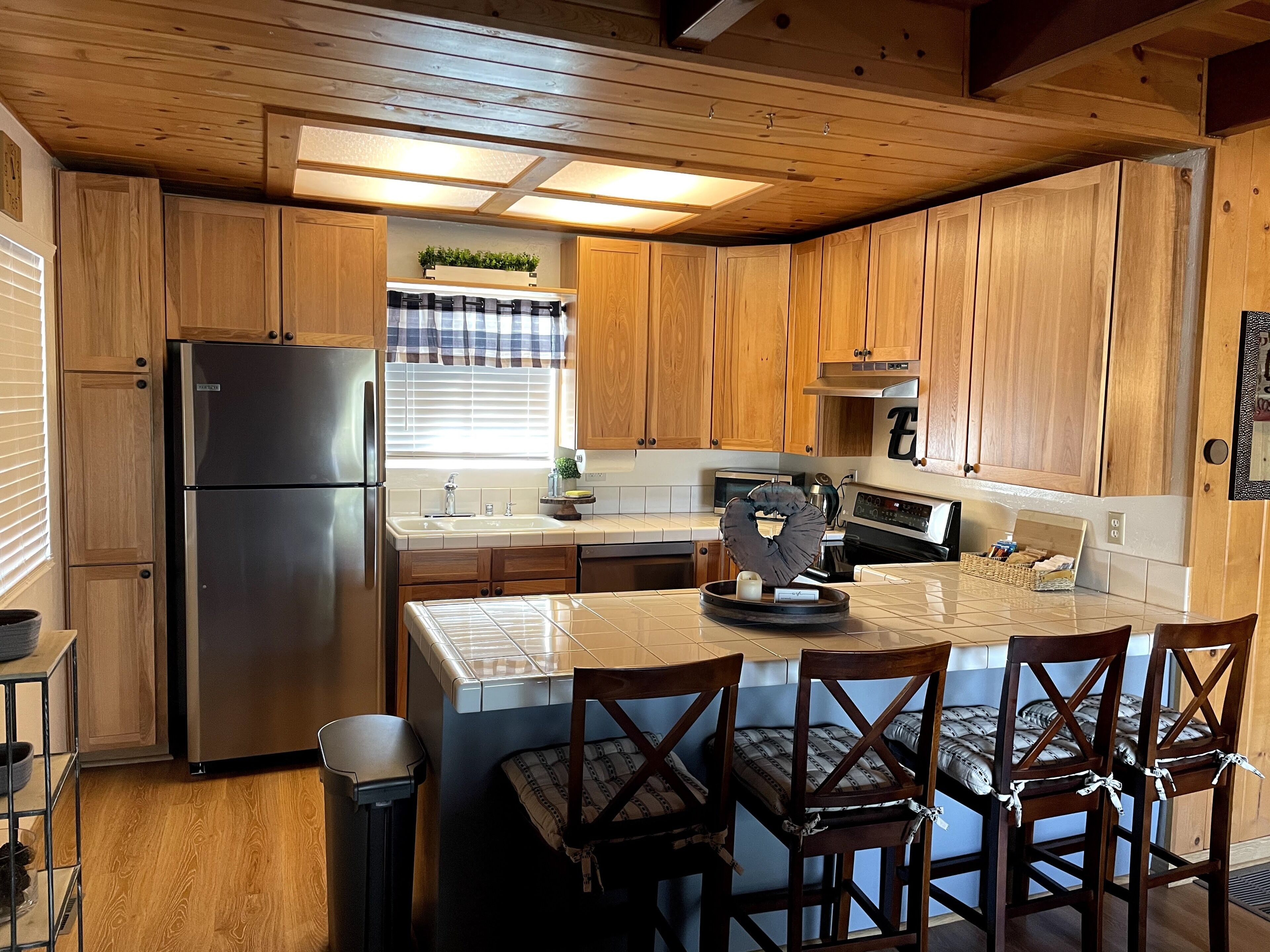 A kitchen with wooden cabinets, a stainless steel refrigerator, a sink, and a stove. There are four wooden chairs around a small island with a kettle and a basket of bread.