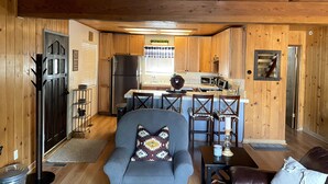 Interior acogedor de una cabaña con paredes de madera, una cocina, una zona de estar con sofá y sillas, y una mesa de comedor.