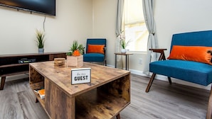 A modern living room with a wooden coffee table, two blue armchairs, a TV mounted on the wall, and a window with curtains. - Quiet Oasis Near the Colorado River STR (Bullhead City)