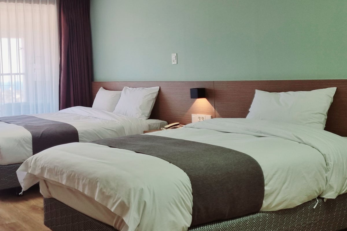 Une chambre d’hôtel avec deux lits, un plancher en bois, une grande fenêtre avec des rideaux légers et une lampe fixée au mur.