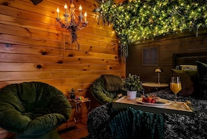 Une pièce chaleureuse aux murs en bois, agrémentée d’un lustre, de chaises vertes, d’une petite table sur laquelle se trouve un plateau avec de la nourriture et des boissons, et d’une plante suspendue illuminée par des lumières.