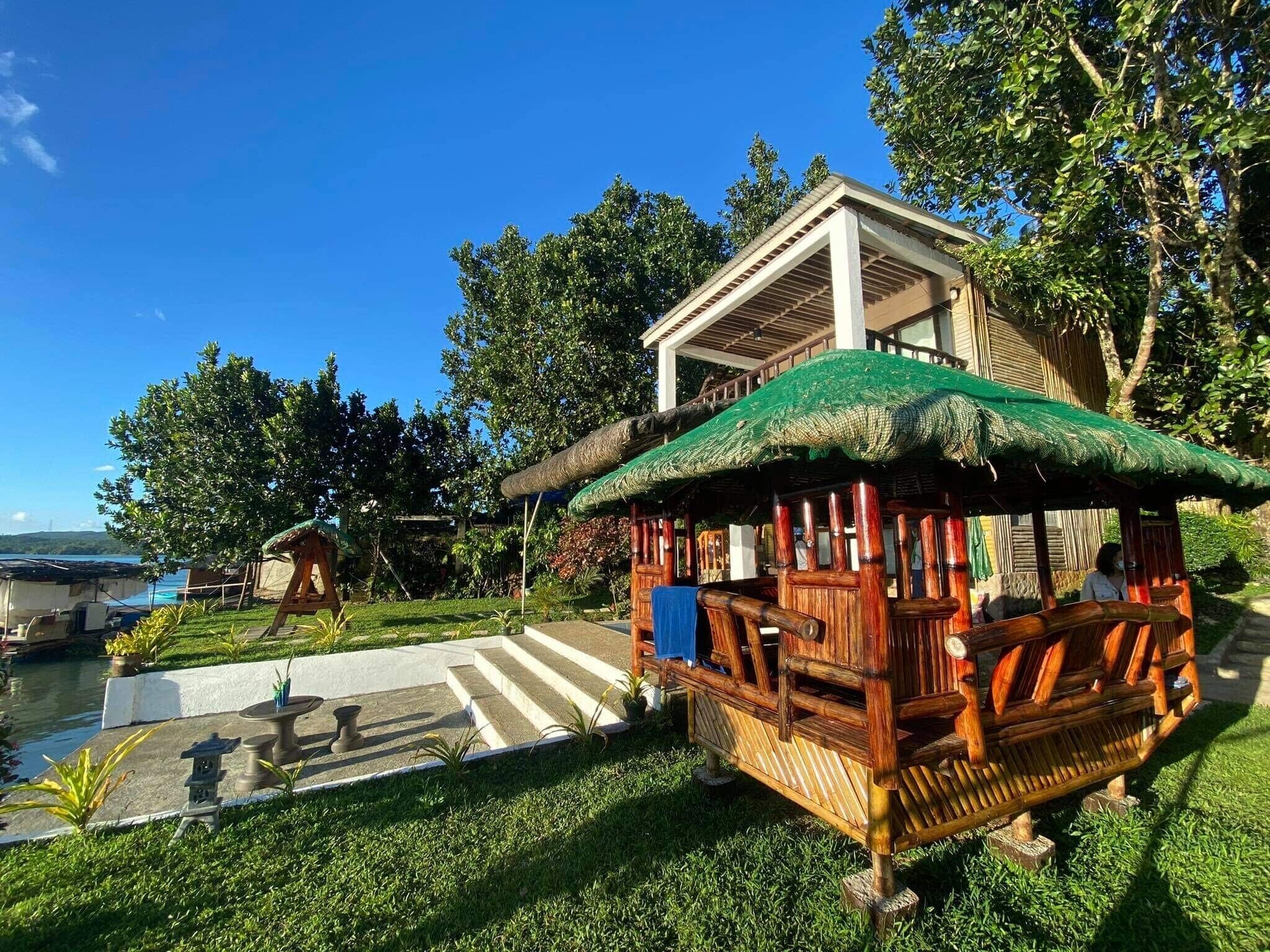 A traditional wooden structure with a thatched roof, situated in a lush garden with a clear blue sky.
