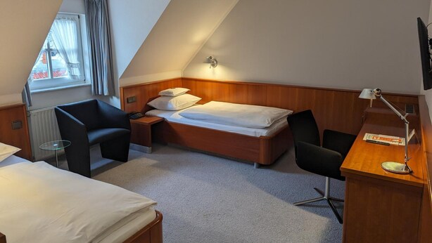 Une chambre d’hôtel avec un plafond en pente, un lit, un bureau avec une lampe, deux chaises et une fenêtre avec des rideaux.