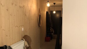 Corredor com paredes de madeira, um radiador e um casaco pendurado sobre ele. Há luminárias suspensas e uma porta que dá para outro quarto.