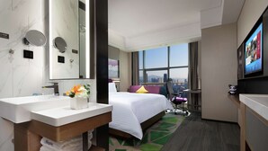 Quarto de hotel com uma cama grande, banheiro com pia e espelho, e televisão na parede.