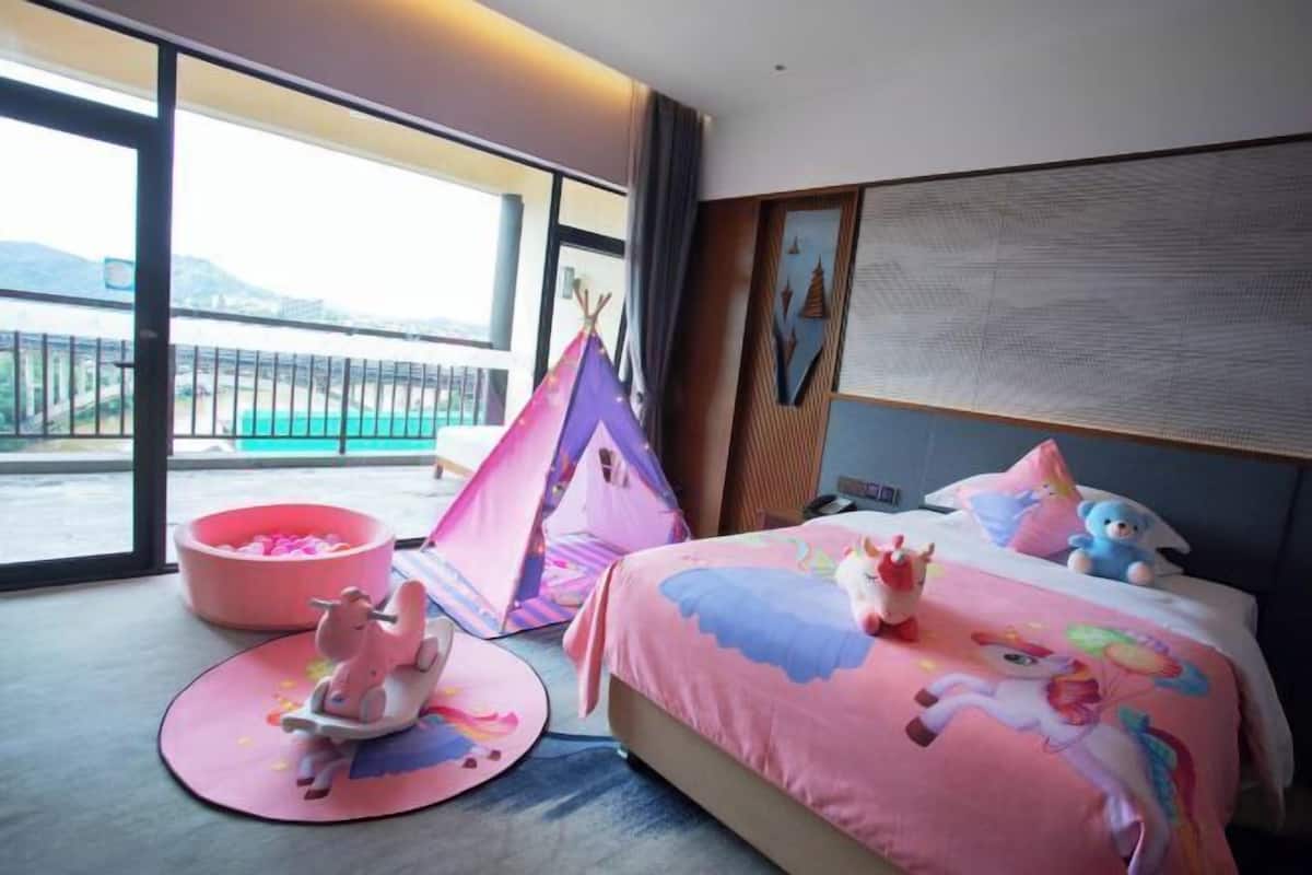 Una habitación de hotel con balcón, una cama con colcha colorida, una zona de juegos rosa y una tienda de campaña.
