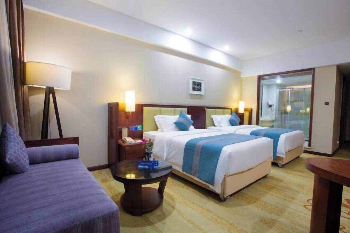 Habitación de hotel con dos camas, un sofá, una mesita y un baño visible a través de una puerta de cristal.