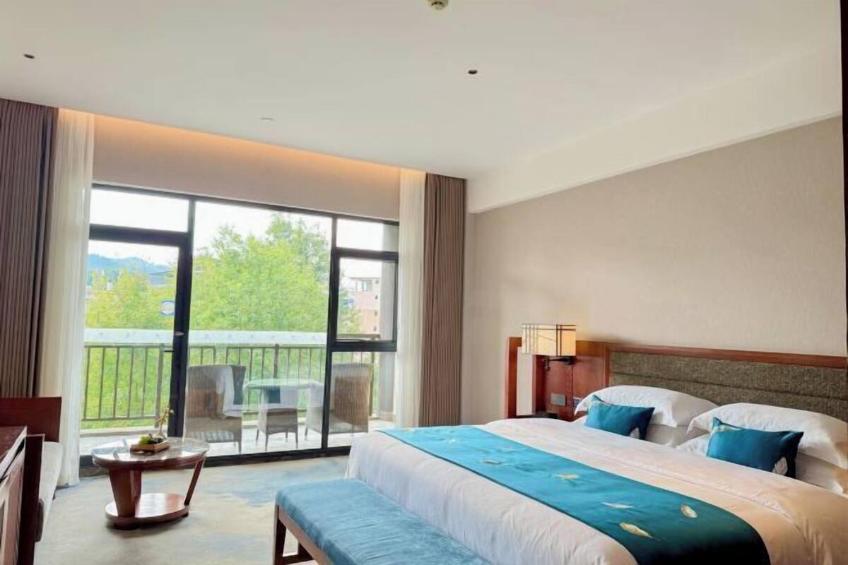 Habitación de hotel con una cama grande, un banco, una mesita y un balcón con vistas a zonas verdes.