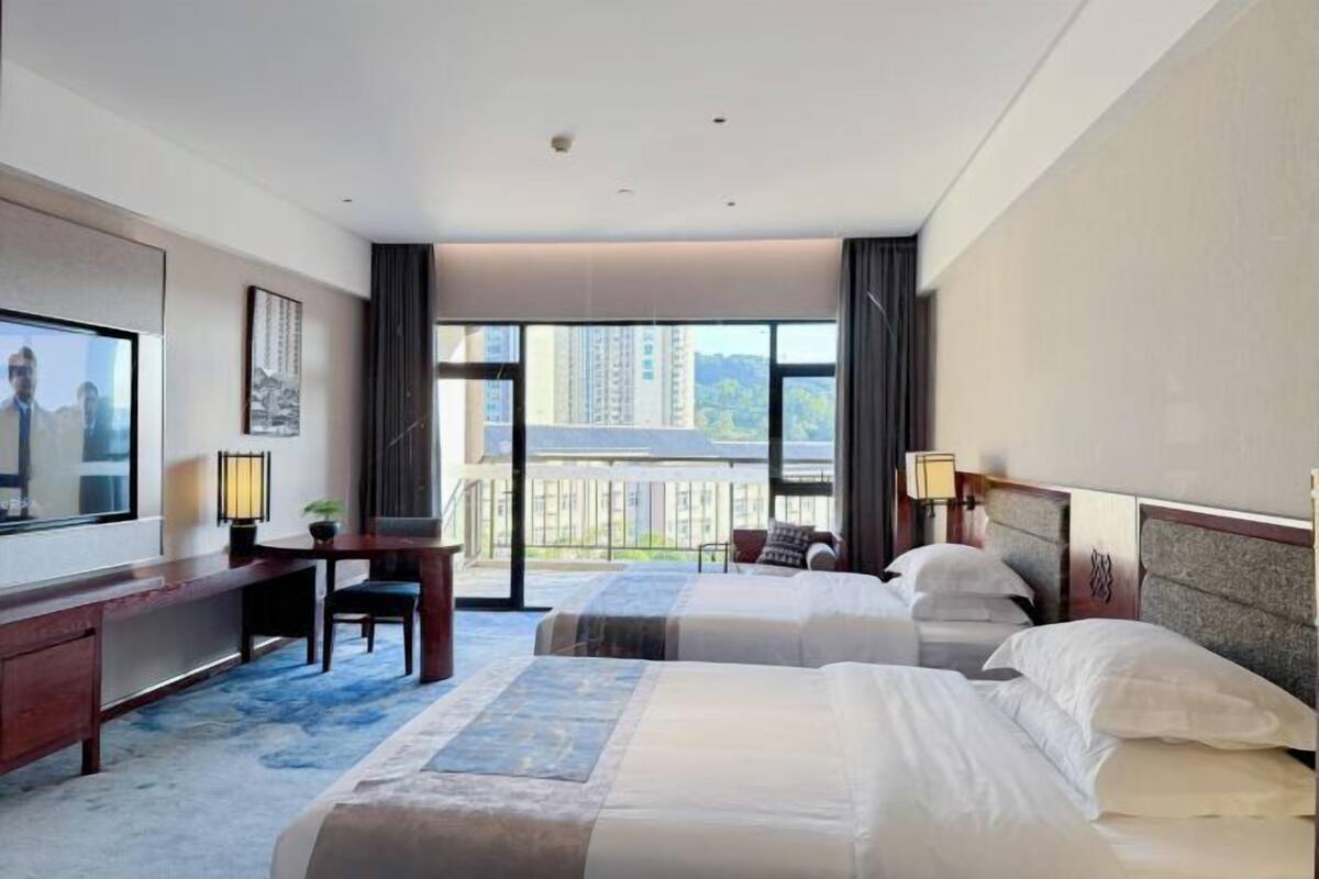 Habitación de hotel con dos camas, un escritorio, una silla, un televisor y un balcón con vista a la ciudad.