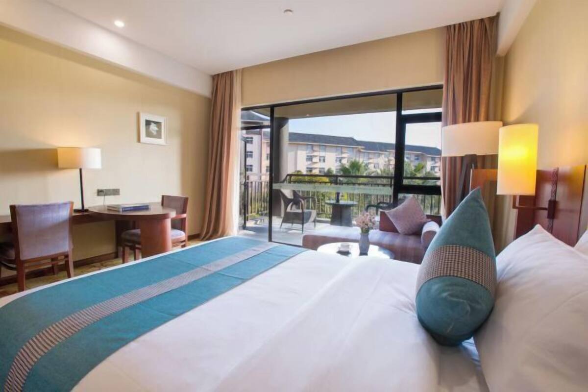 Habitación de hotel con una cama grande, un escritorio, una silla, un balcón con vistas a edificios y una ventana con cortinas.