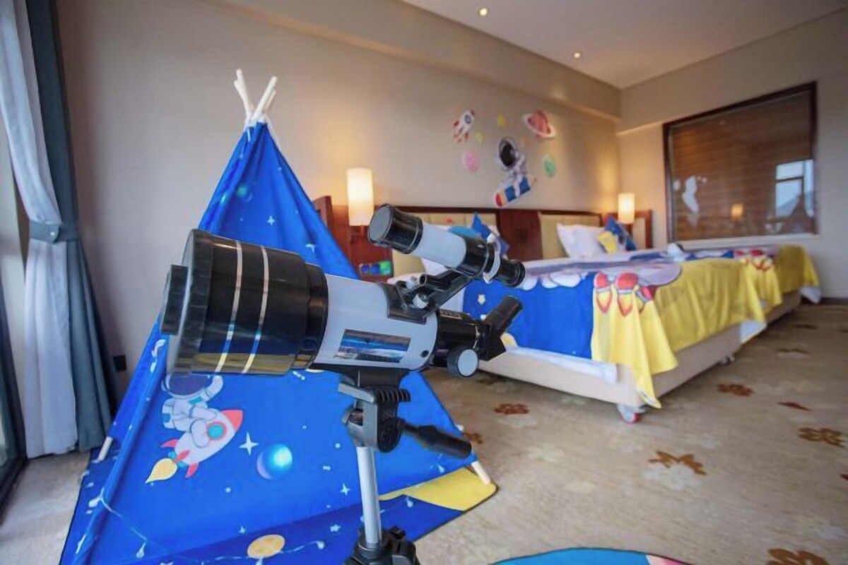 A hotel room with a telescope, a bed with a yellow blanket, and a window with blinds.