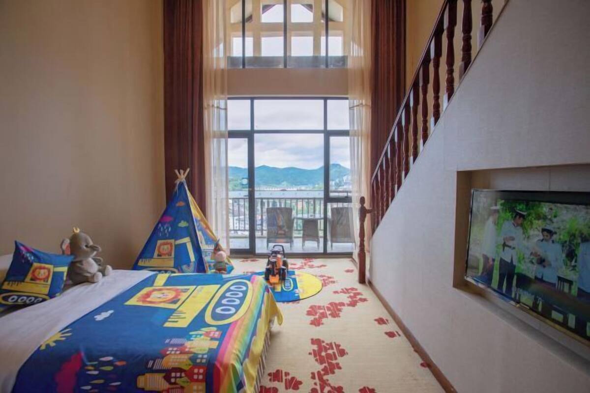 Habitación de hotel con un ventanal grande, una cama con colcha colorida, una zona de juegos pequeña con un tipi y un televisor en la pared.