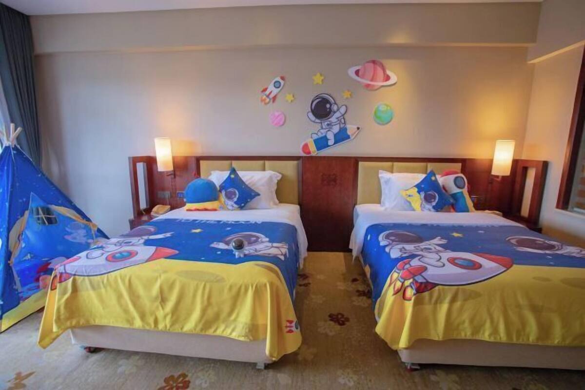 Habitación de hotel con dos camas, cada una con cubrecama amarillo y almohadas azules, una estructura azul similar a una tienda de campaña y decoraciones en la pared, incluyendo un astronauta de dibujos animados.