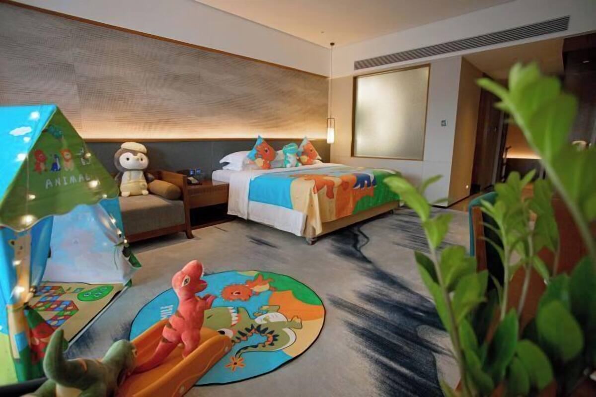 Una habitación de hotel con una cama, una mesita de noche, una lámpara, una casa de juguete y un dinosaurio de juguete en el suelo.