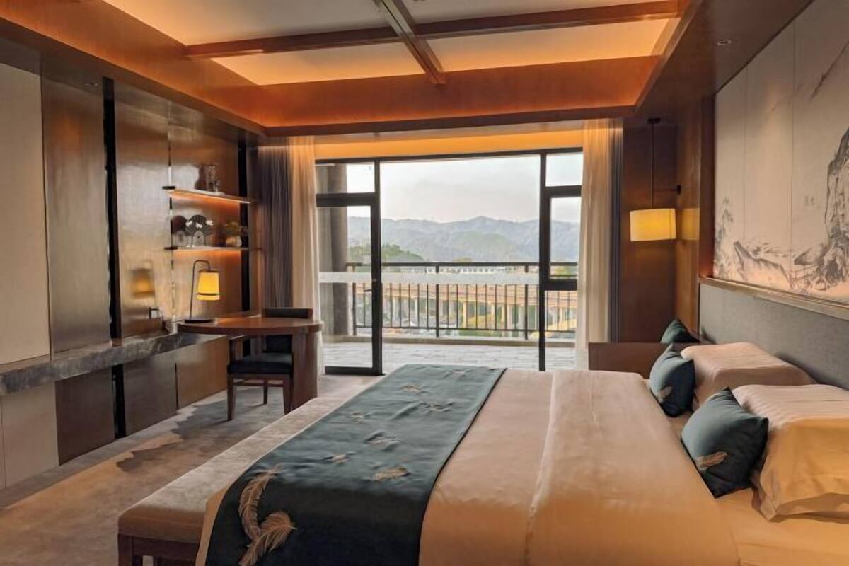 Habitación de hotel con una cama grande, un escritorio y vistas a montañas desde un balcón.