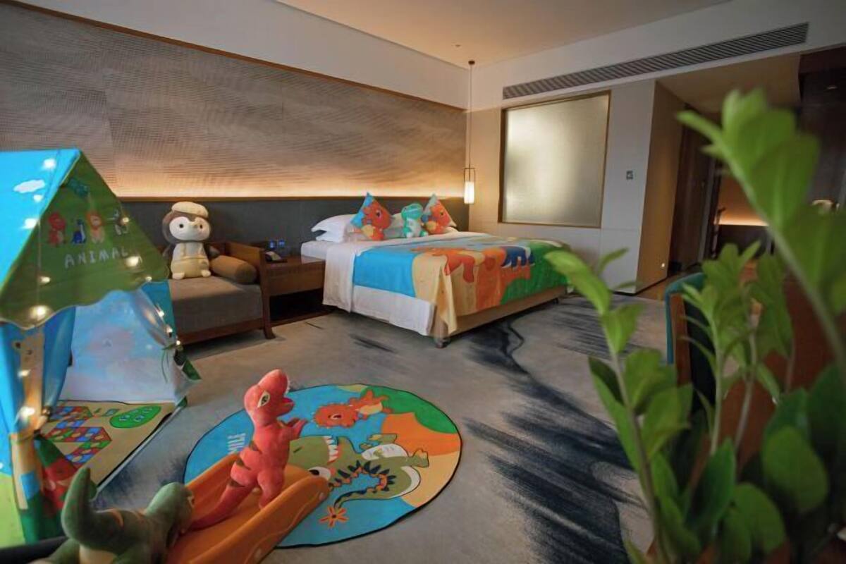 Habitación de hotel con cama, mesita de noche, lámpara, ventana y un área de juegos infantil con una tienda de campaña y juguetes.