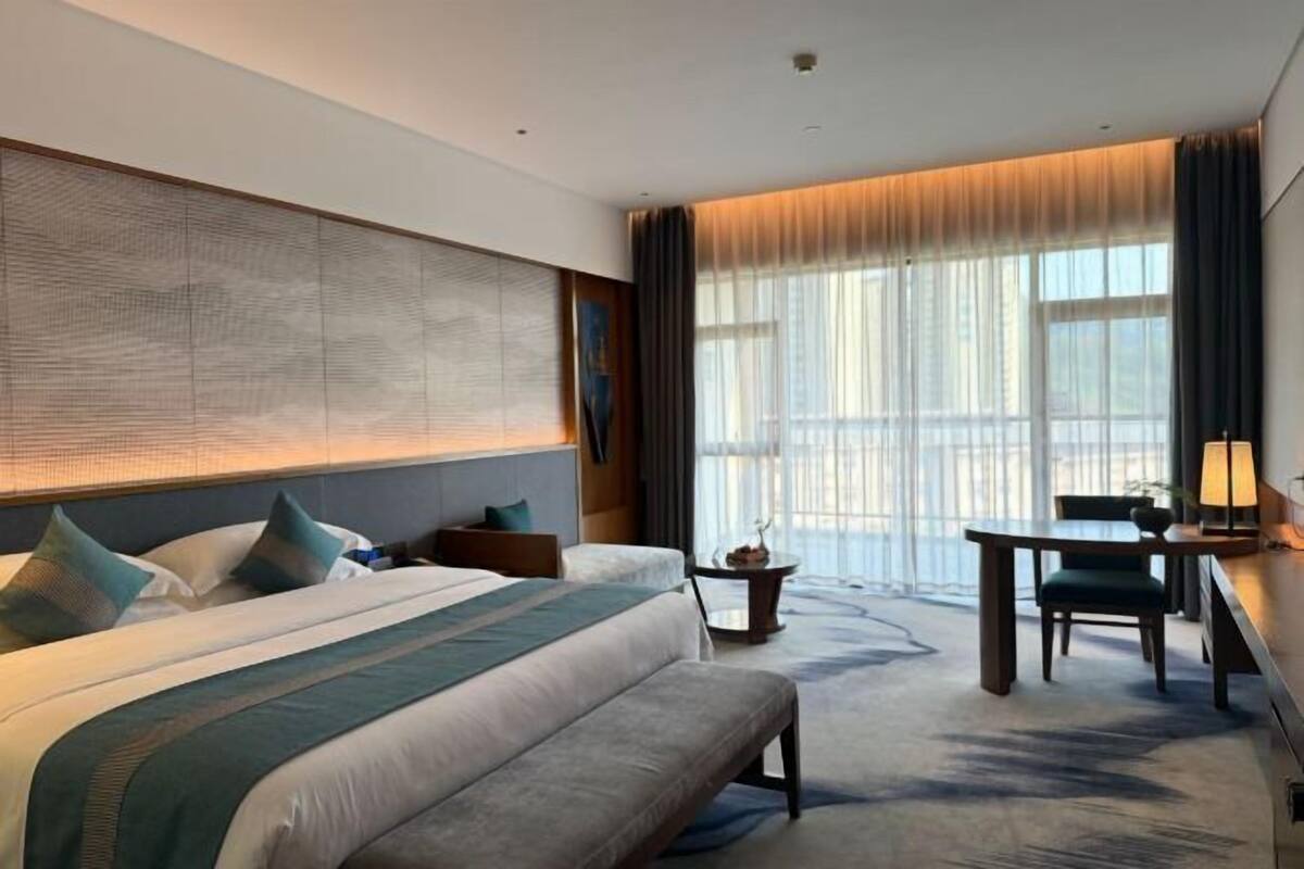 Habitación de hotel con una cama grande, un banco, un escritorio y vistas de la ciudad a través de amplios ventanales.