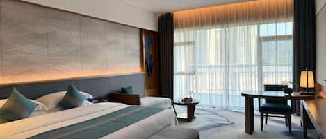 A hotel room with a large bed, a bench, a desk, and a view of the city through large windows.