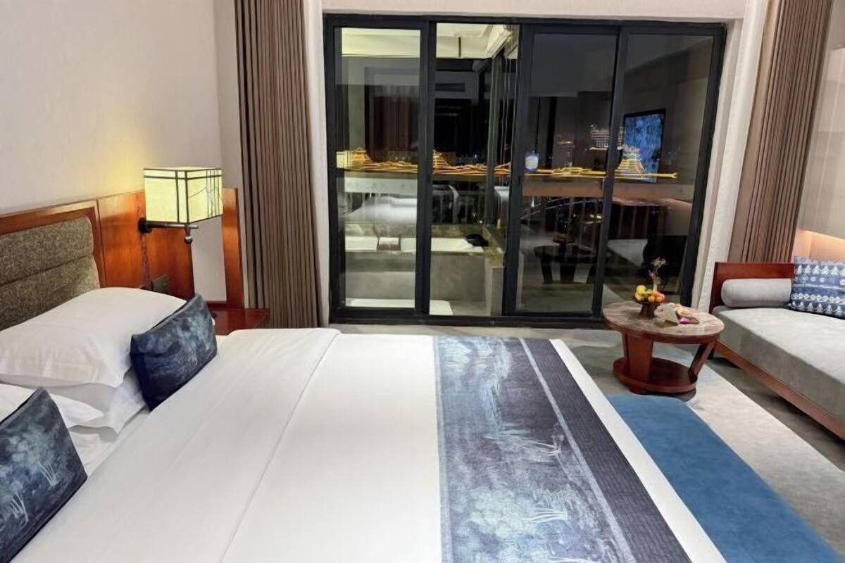 Habitación de hotel con una cama grande, una mesita redonda con un florero y vista a un comedor por una puerta de vidrio.