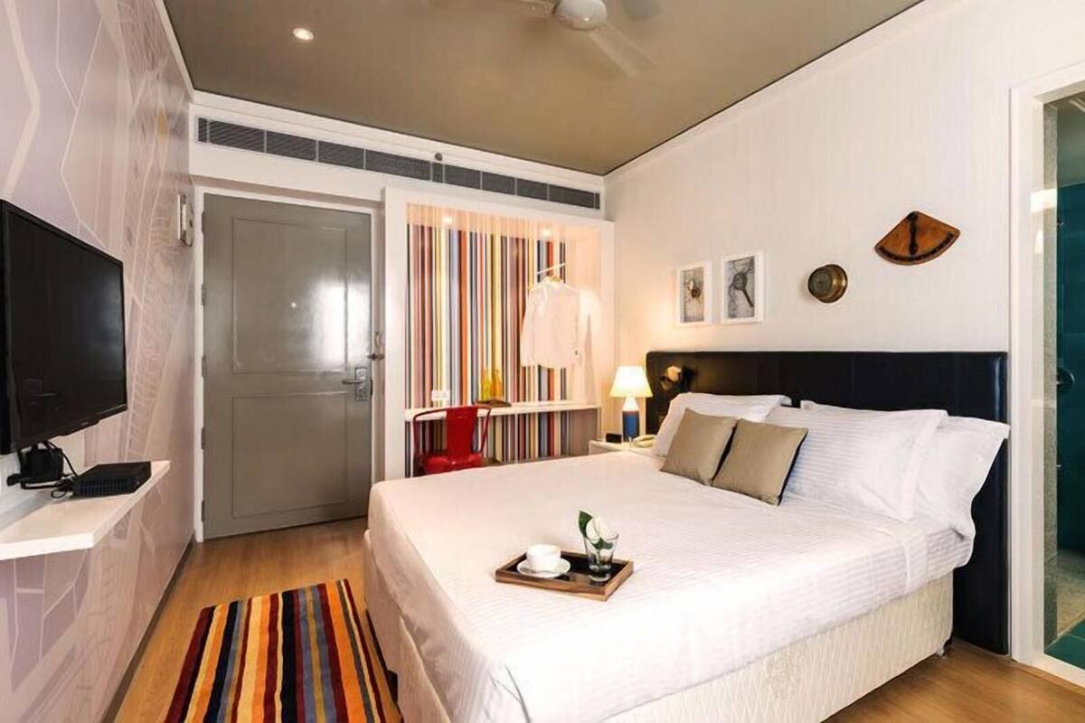 Habitación de hotel con cama, televisión, escritorio con computadora, estantería y baño visible a través de una puerta abierta.