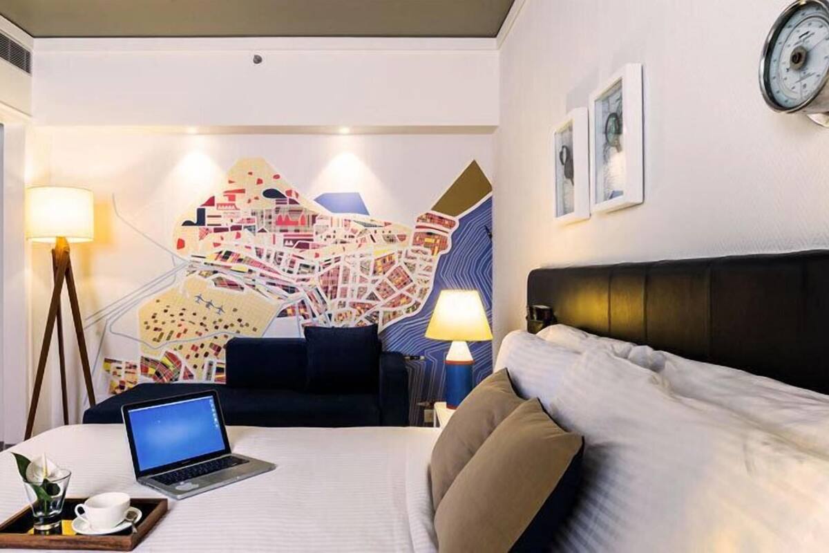 Habitación de hotel con cama, escritorio con ordenador portátil, mapa en la pared y dos lámparas.