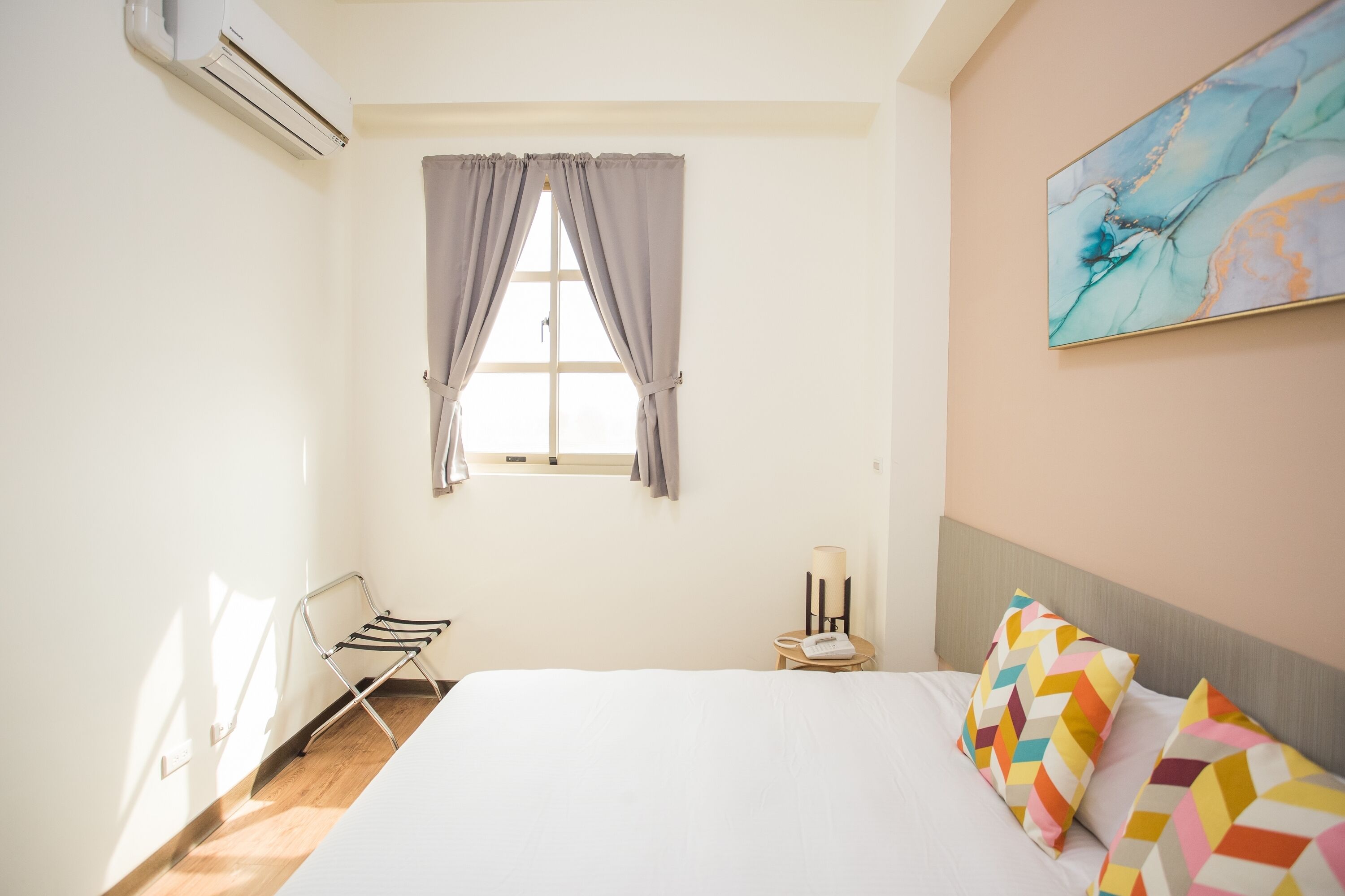 Habitación de hotel con cama, ventana con cortinas, aire acondicionado de pared, mesita con lámpara y un cuadro de colores en la pared.