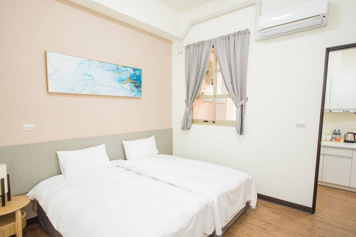A hotel room with a bed, a window with curtains, a painting on the wall, and a kitchenette.