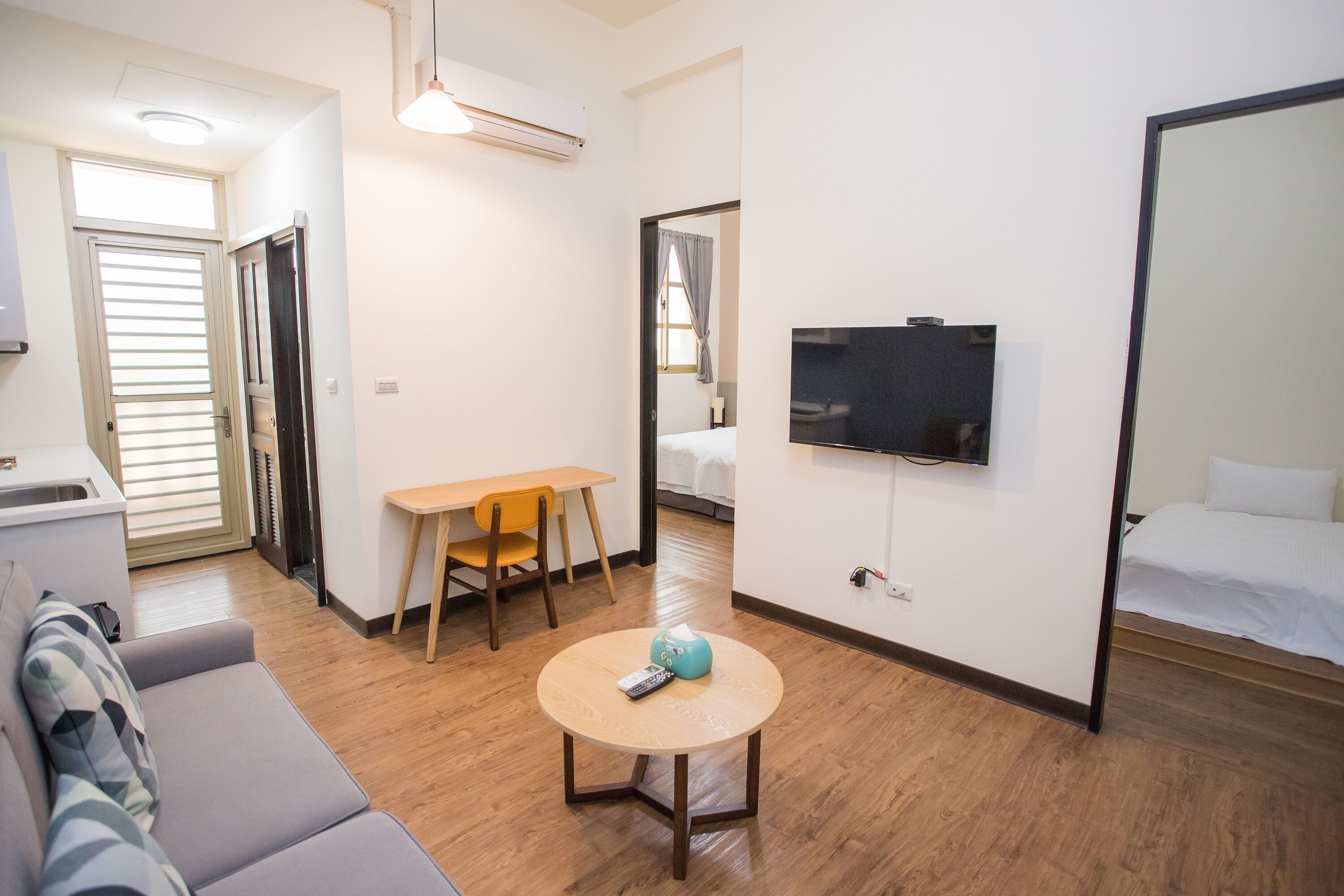 Habitación de hotel con sofá, una pequeña mesa redonda con un control remoto, televisor montado en la pared, cama y una puerta que da a otra habitación.