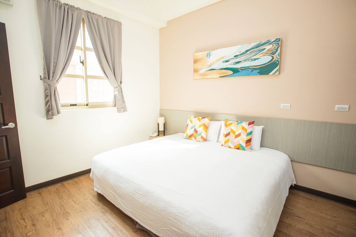 A hotel room with a large bed, a window with curtains, a painting on the wall, and a nightstand with a lamp.