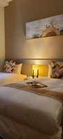 Habitación de hotel con dos camas, una mesita de noche con lámpara, un cuadro en la pared y una bandeja con taza y platillo sobre la cama.