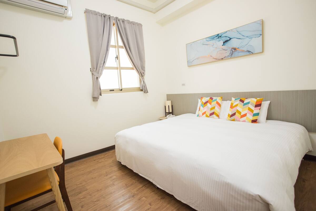 A hotel room with a bed, a desk, a chair, a window with curtains, and a painting on the wall.