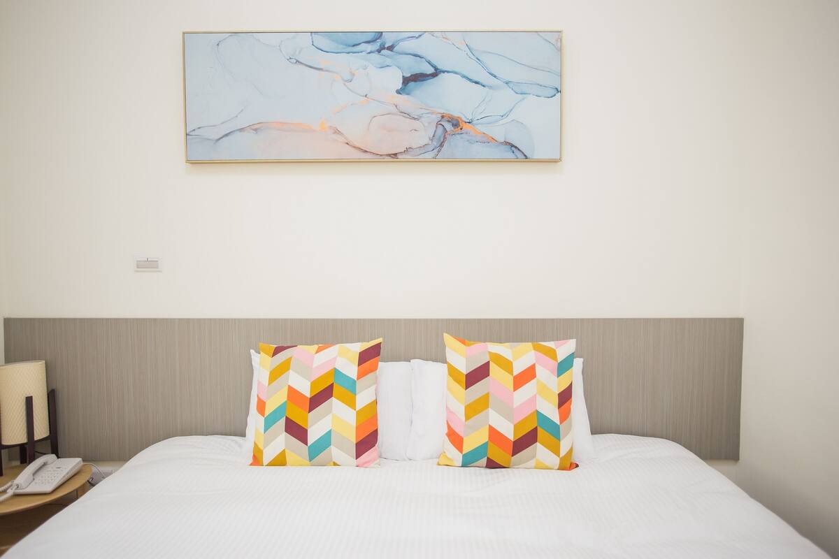 A hotel room with a large abstract painting on the wall, a bed with white linens, two colorful chevron-patterned pillows, a bedside lamp, and a phone on the nightstand.