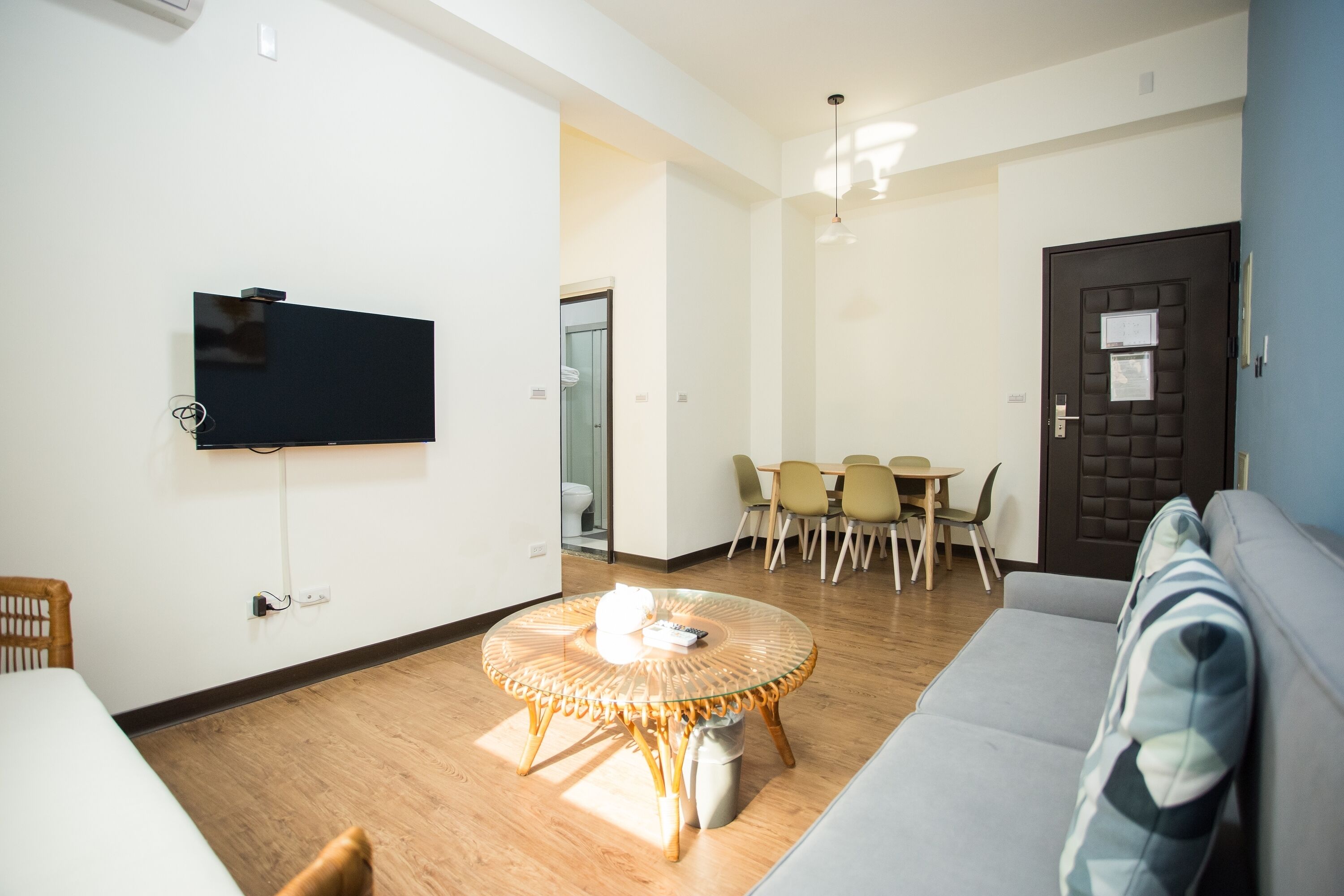 A modern living room with a flat-screen TV mounted on the wall, a round glass table with a wicker base, a grey sofa, and a dining area with a table and chairs.