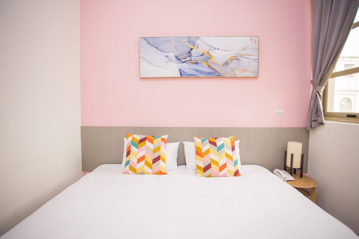 A hotel room with a bed, two decorative pillows, a nightstand, a lamp, and a painting on the wall.