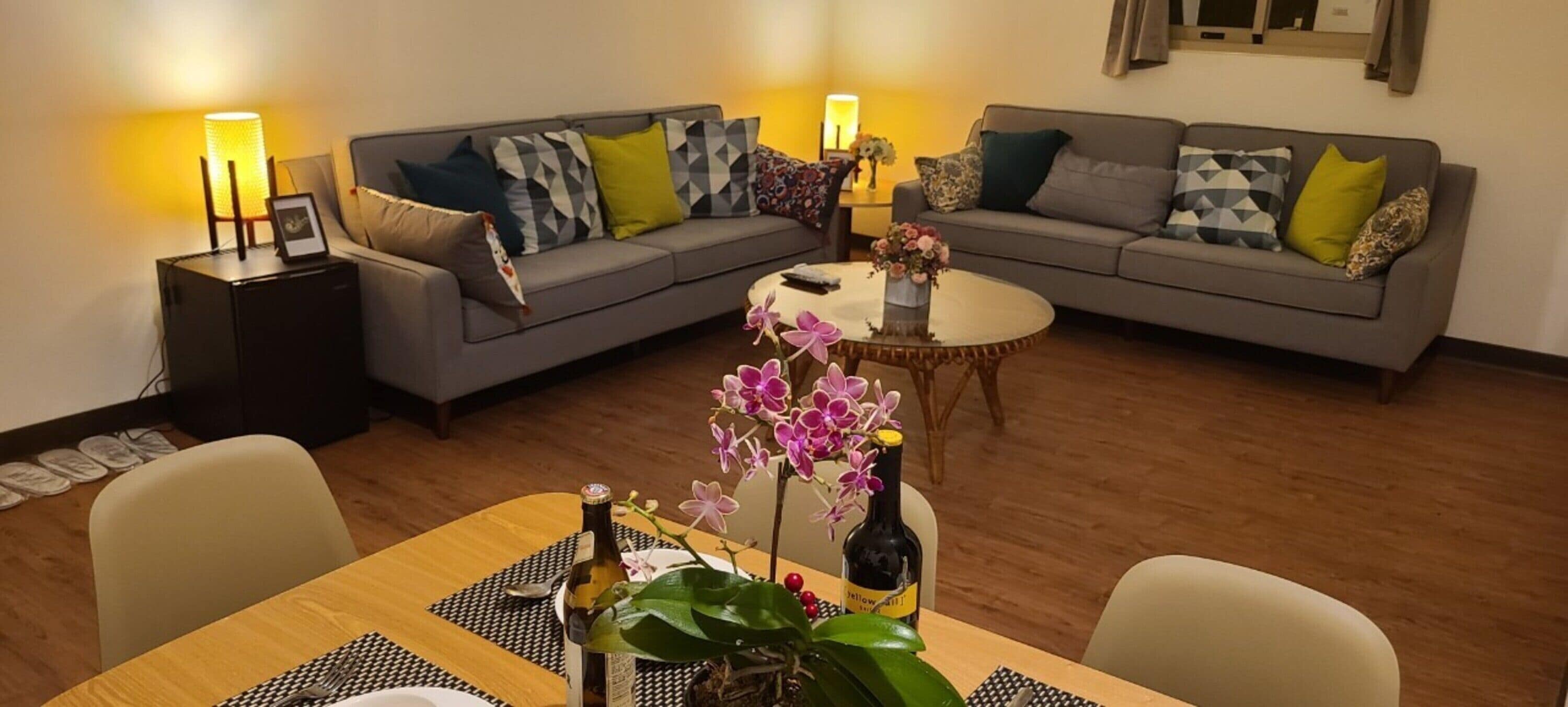 A living room with two grey sofas, a round coffee table, and a dining table with chairs. There are lamps on side tables, a bottle of wine, and a vase with flowers.