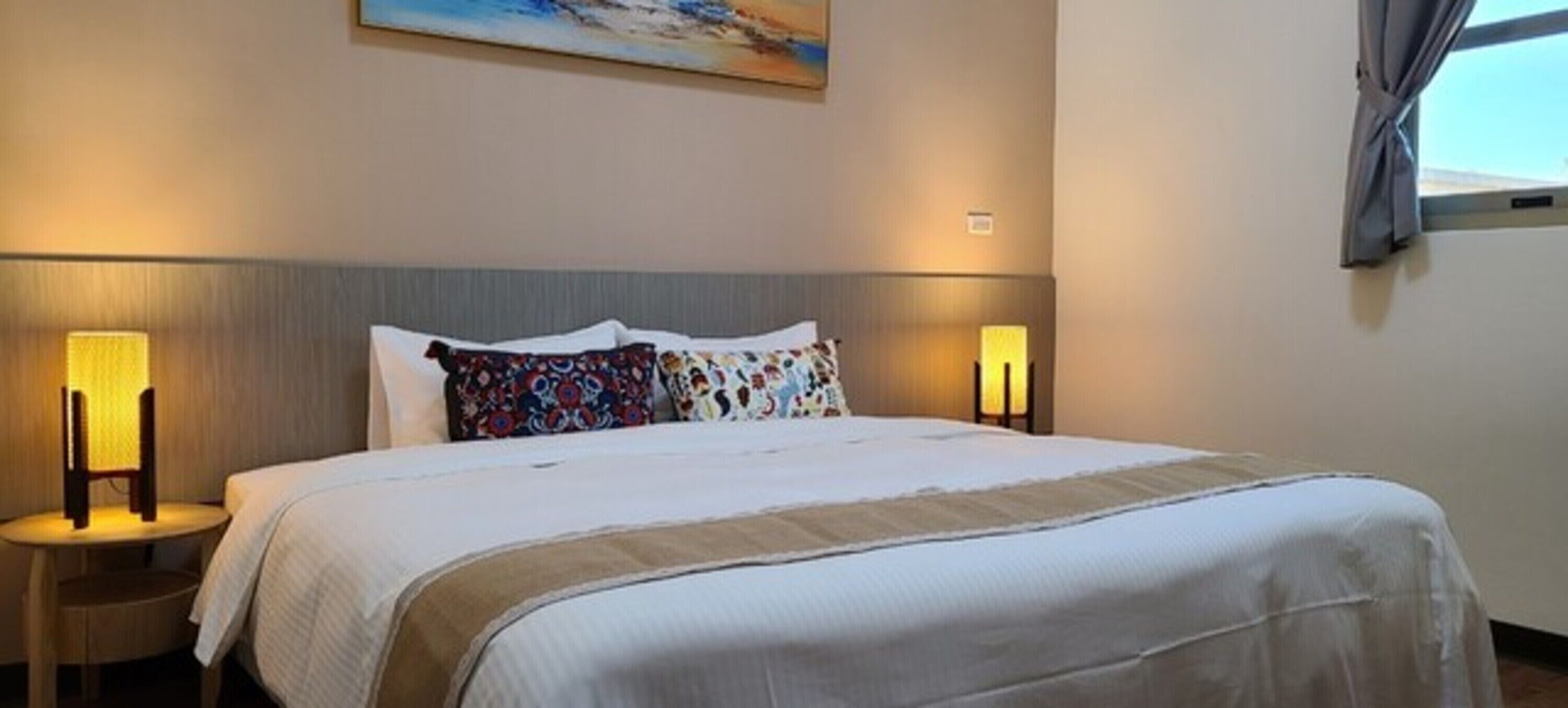 A hotel room with a large bed, two bedside lamps, a headboard, and a window with curtains.