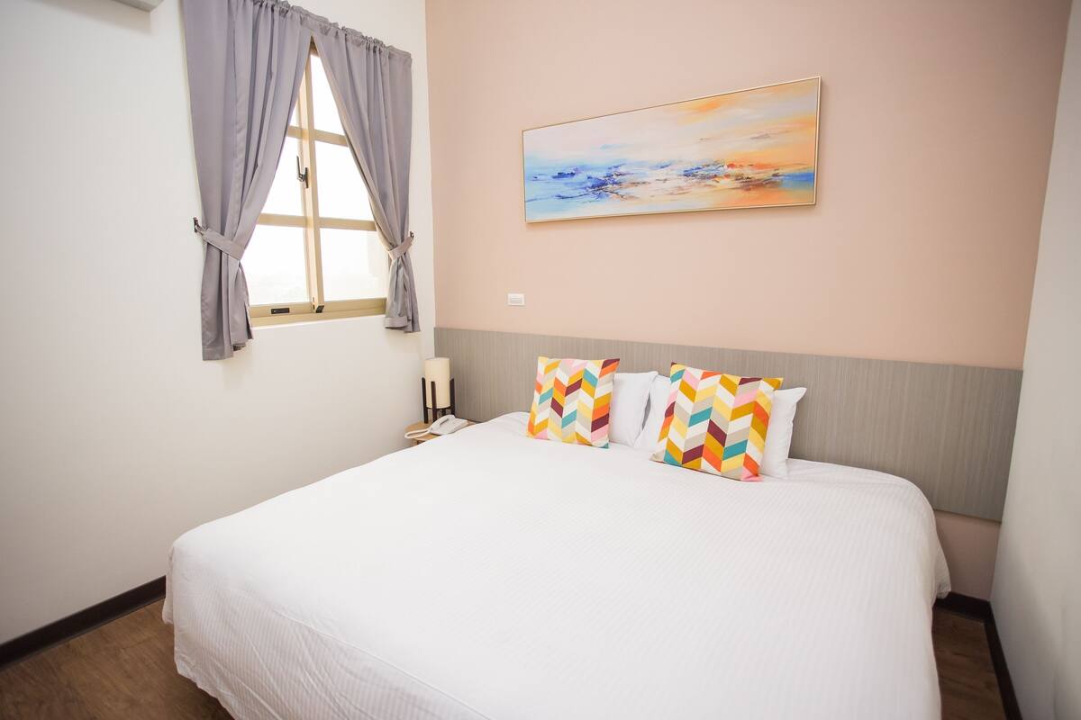 A hotel room with a large bed, a window with curtains, a painting on the wall, and a nightstand with a lamp and phone.