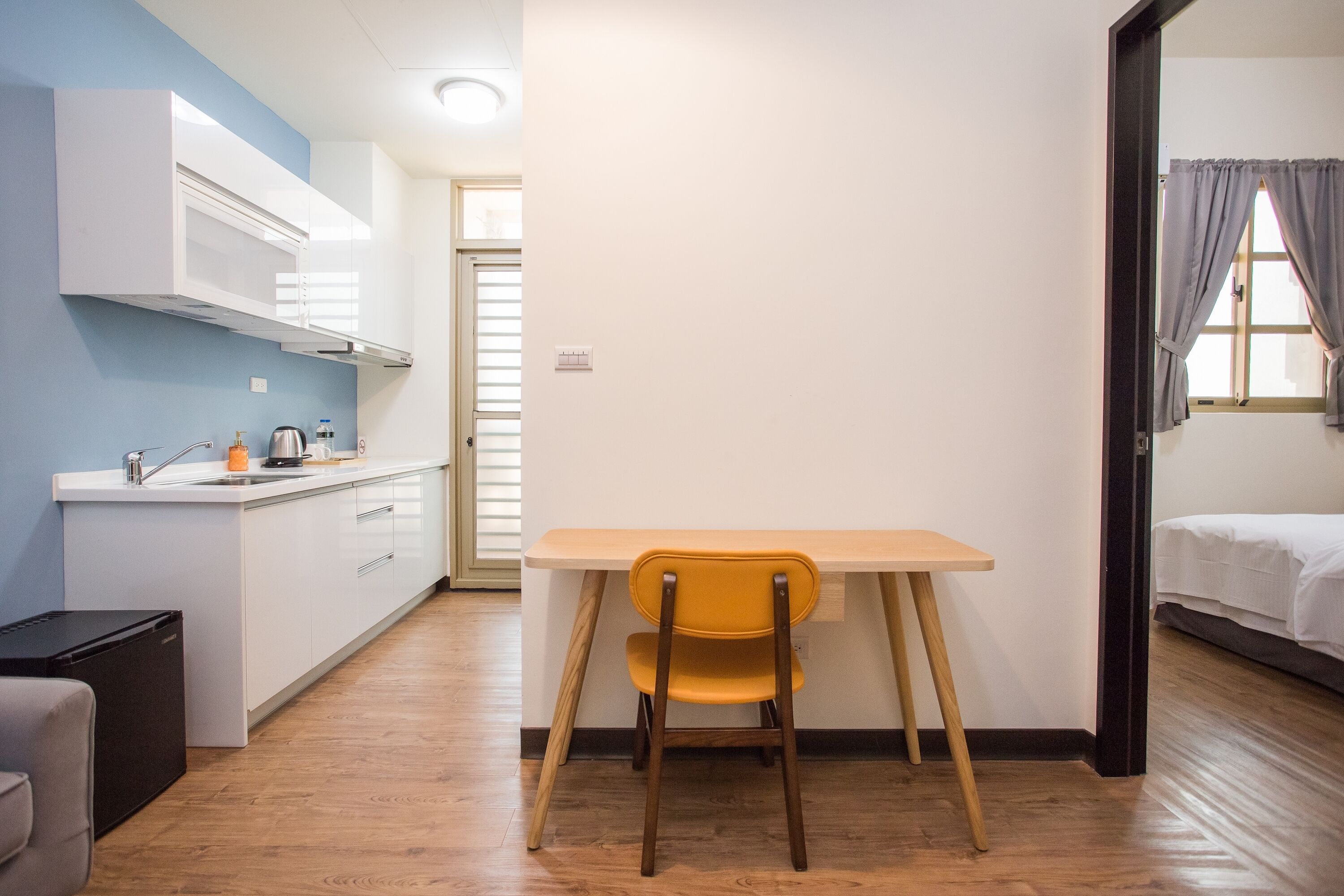 A compact kitchen with white cabinets, a small refrigerator, and a wooden dining table with a yellow chair.