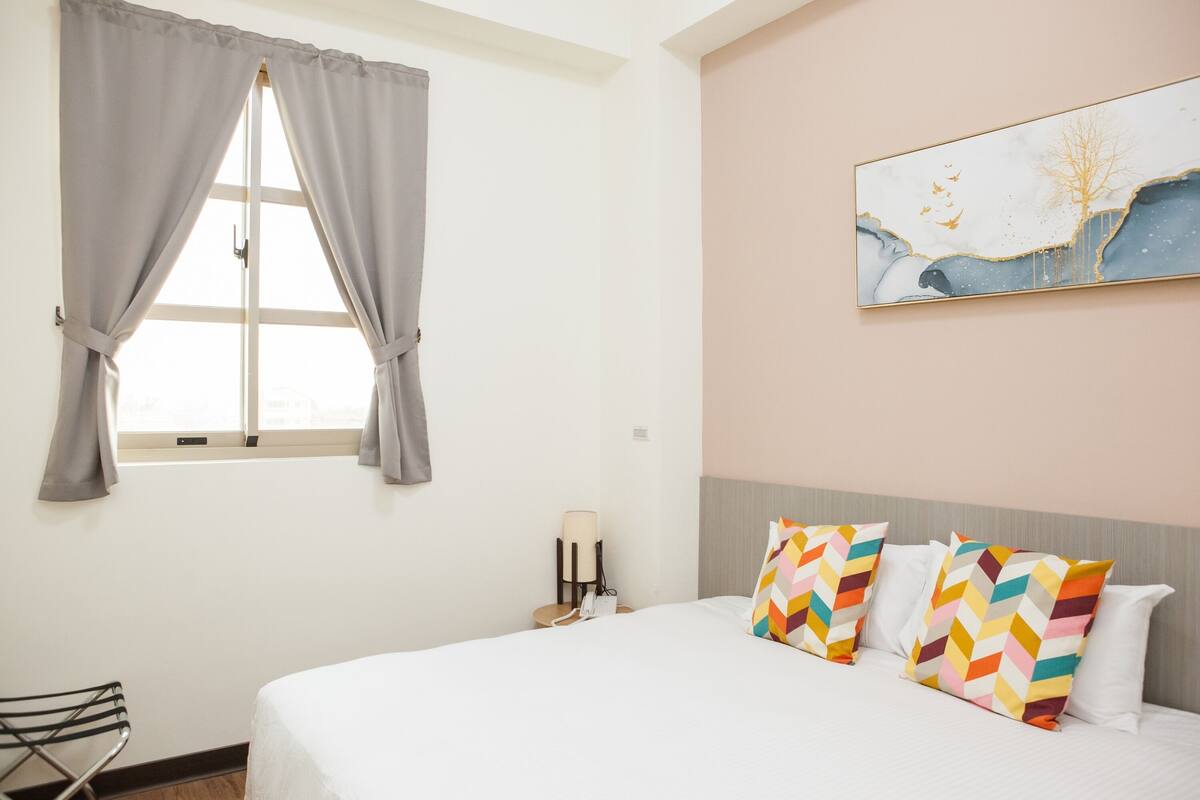 A hotel room with a bed, a window with curtains, a wall-mounted lamp, and a painting.