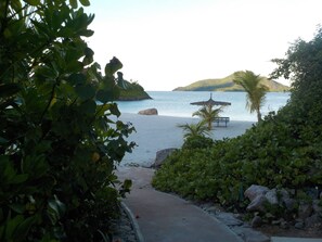 A beach pathway leading to a bench and umbrella by the sea, surrounded by greenery and rocks.