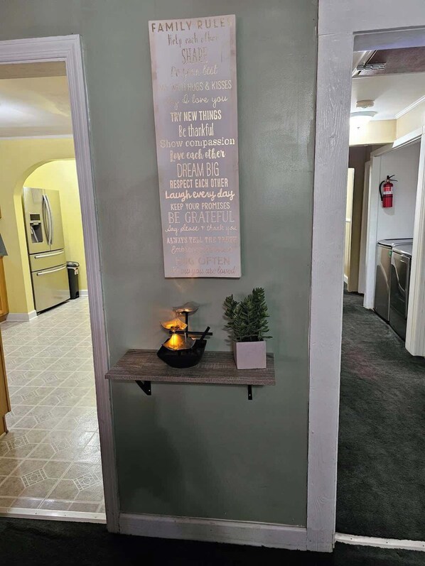 Een gang met een bord aan de muur waarop familieregels staan, een klein tafeltje met een kaars en een potplant.